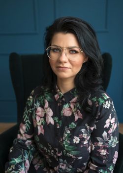 Adwokat Marta Szymańczuk, profesjonalny portret w biurze z tłem w odcieniu ciemnoniebieskim, ubrana w kwiecistą koszulę.